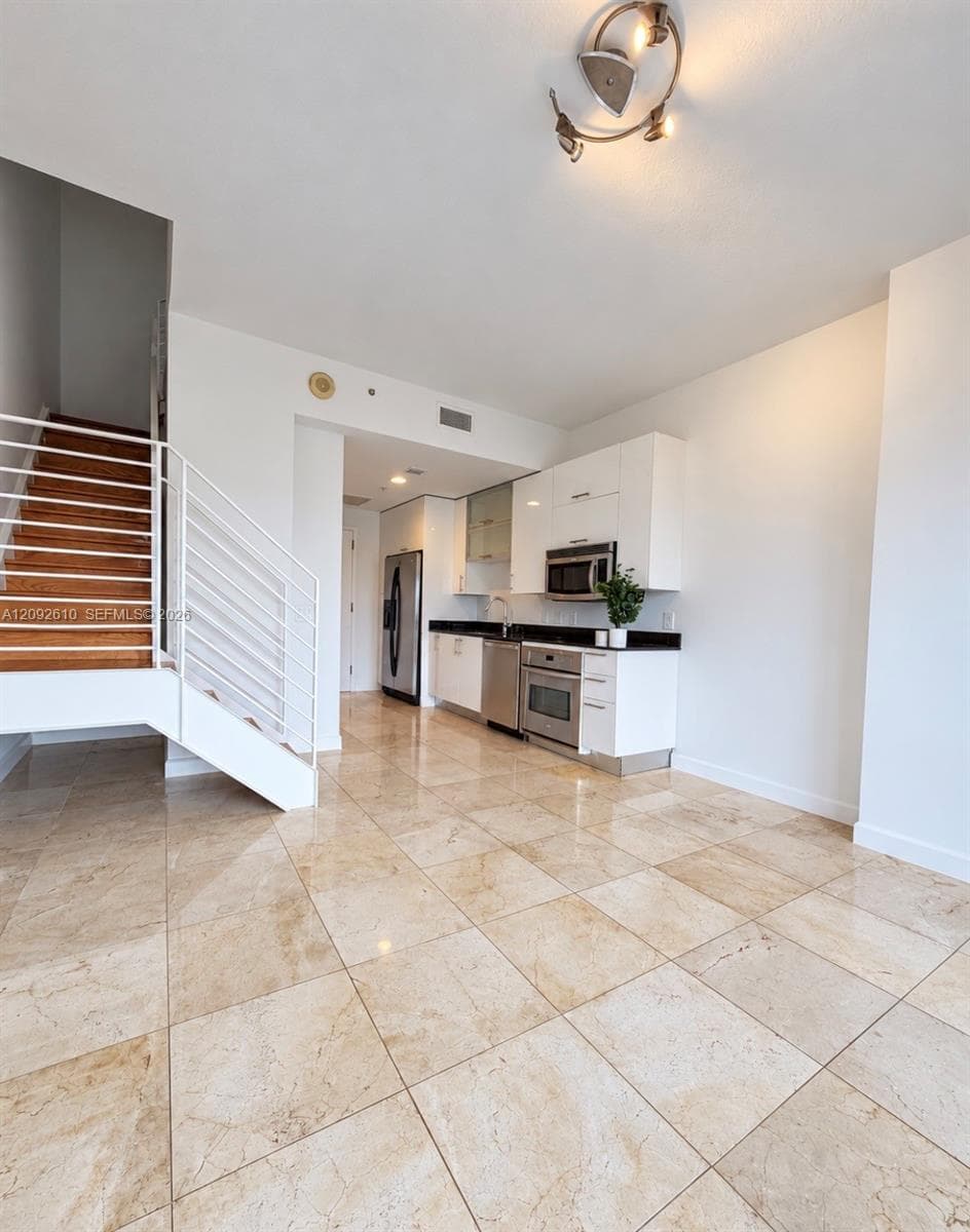 Modern kitchen featuring white flat-panel cabinetry, dark stone countertops, stainless steel appliances, polished stone flooring, and a staircase with a metal railing