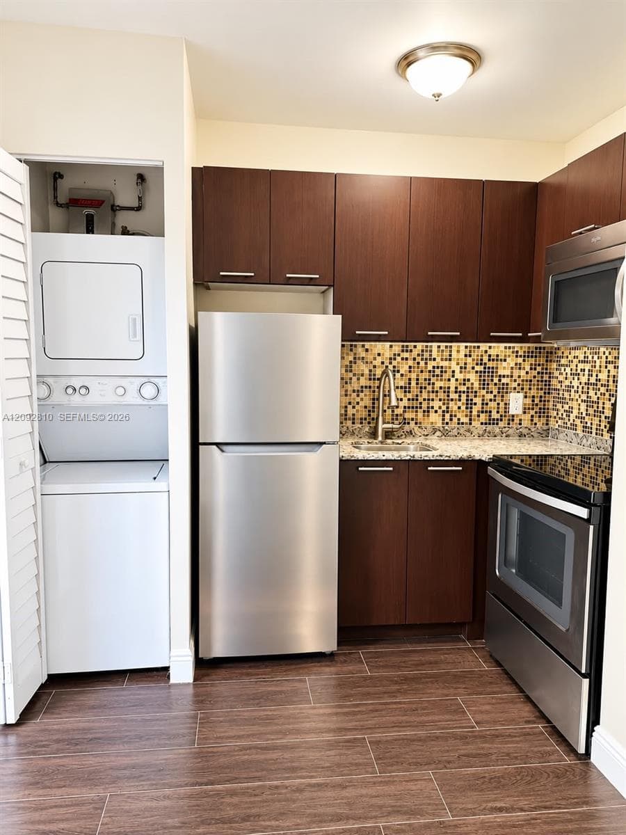 Kitchen featuring dark wood-finish cabinetry, stainless steel appliances, speckled stone-finish countertops, mosaic tile backsplash, and wood-finish flooring with an integrated laundry closet