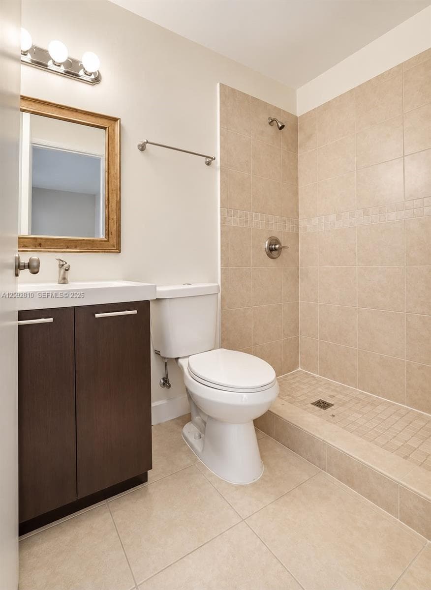 Bathroom featuring a vanity with dark wood-finish cabinetry, a white basin, neutral-toned tile flooring, and a walk-in shower with tiled walls and a mosaic tile accent border