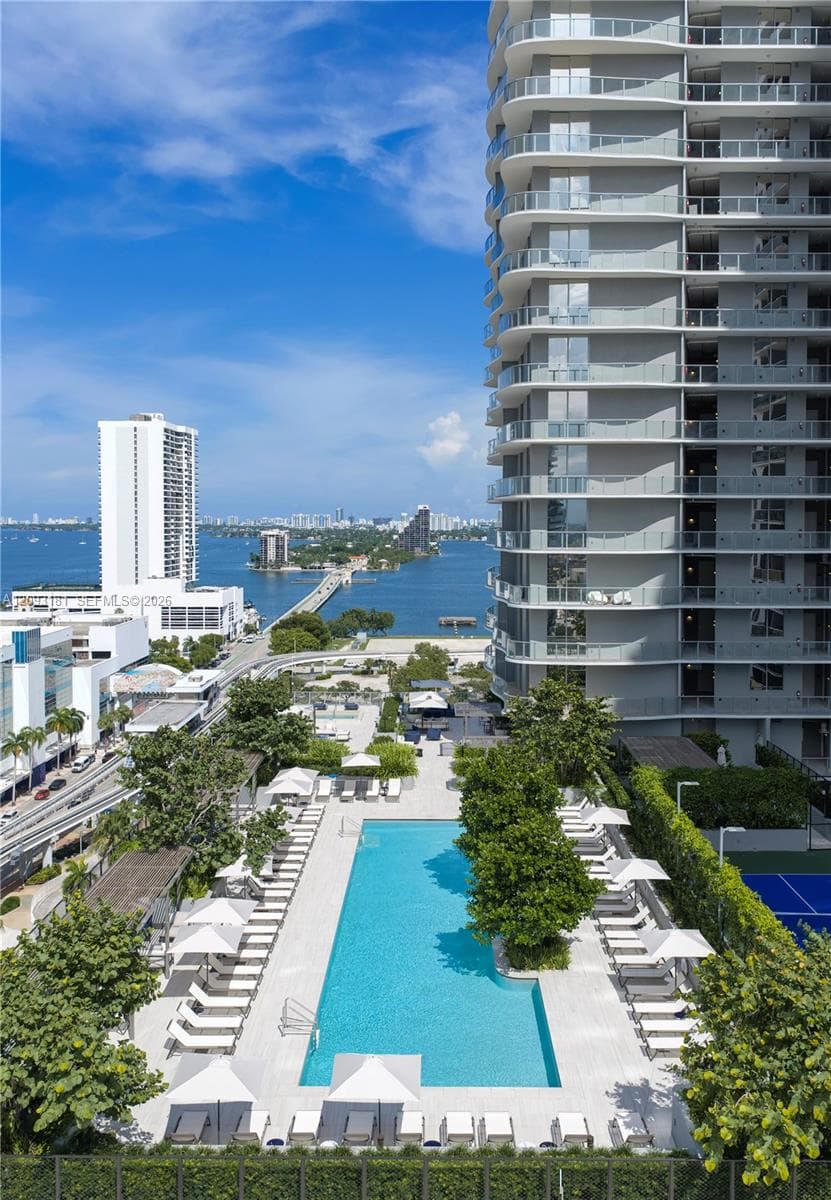 High-rise residential exterior featuring stacked private balconies, rectangular swimming pool with paved terrace, mature landscaping, and hard surface athletic court