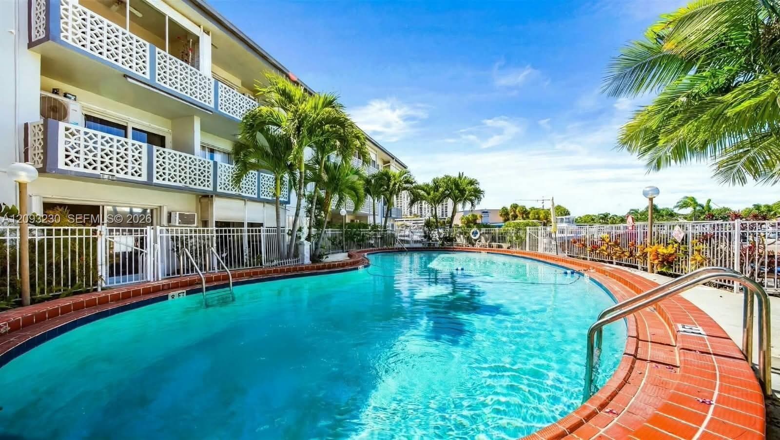 Curved outdoor pool with a brick coping, surrounded by a white metal fence and palm trees