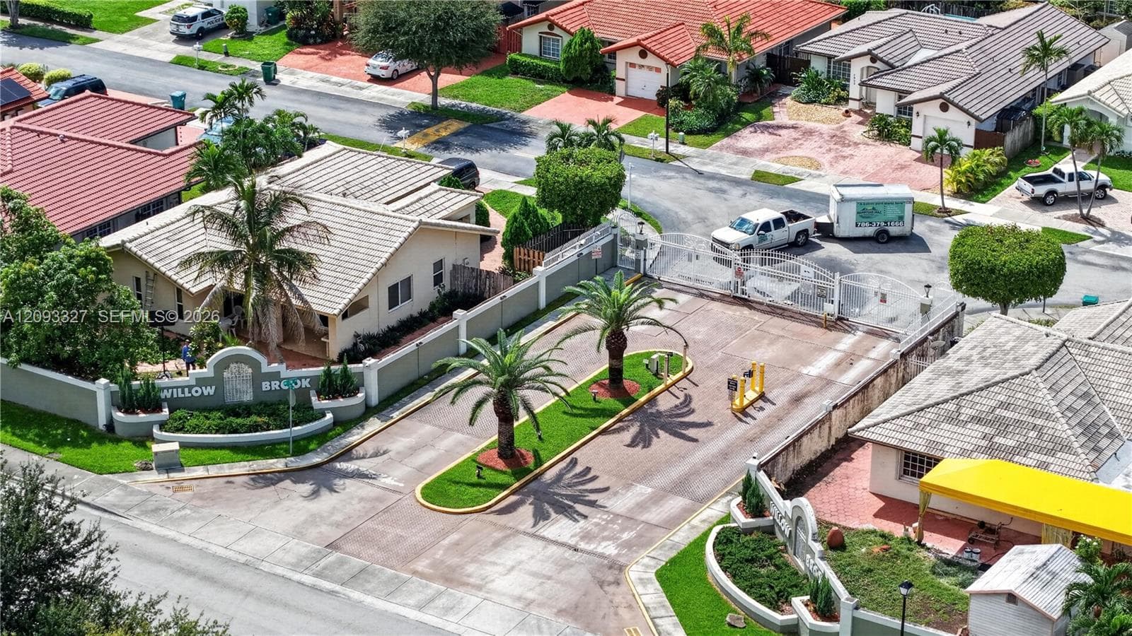 Gated community entrance featuring a secure perimeter wall, decorative signage, paved drive, manicured landscaping, and palm trees