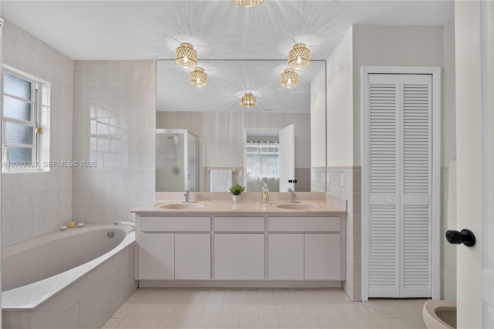 Double vanity featuring dual integrated sinks, large wall-to-wall mirror, built-in soaking tub, tile surround, and louvered closet doors