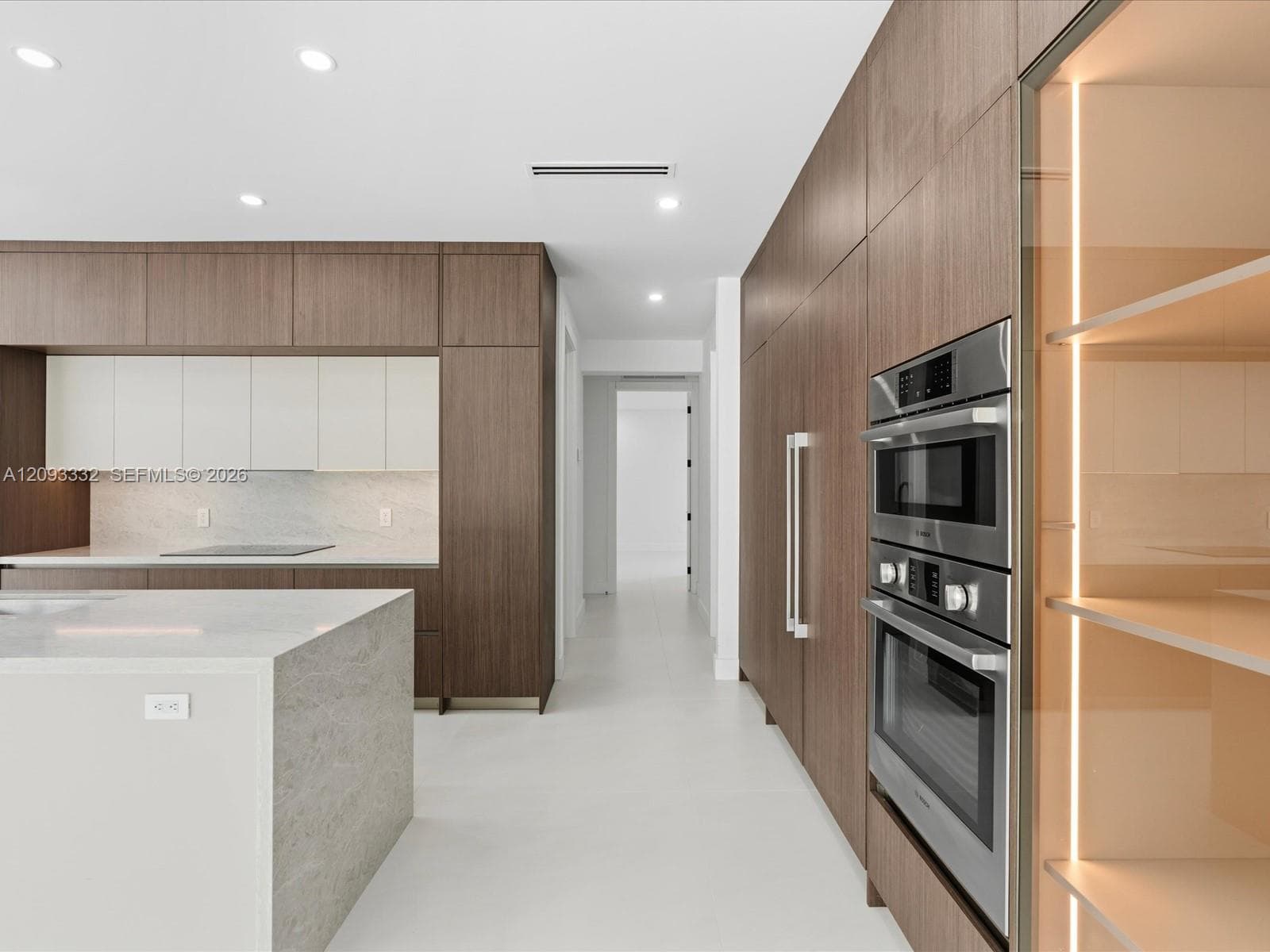 Modern kitchen featuring full-height wood-finish cabinetry, a stone-surfaced waterfall island, integrated stainless steel double wall ovens, and illuminated glass display shelving