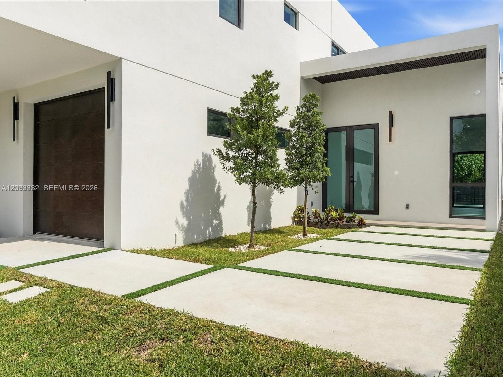Modern exterior featuring a smooth stucco facade, a dark-finished panel garage door, architectural linear wall-mounted lighting, a paved driveway with grass inserts, and double glass-panel entry doors