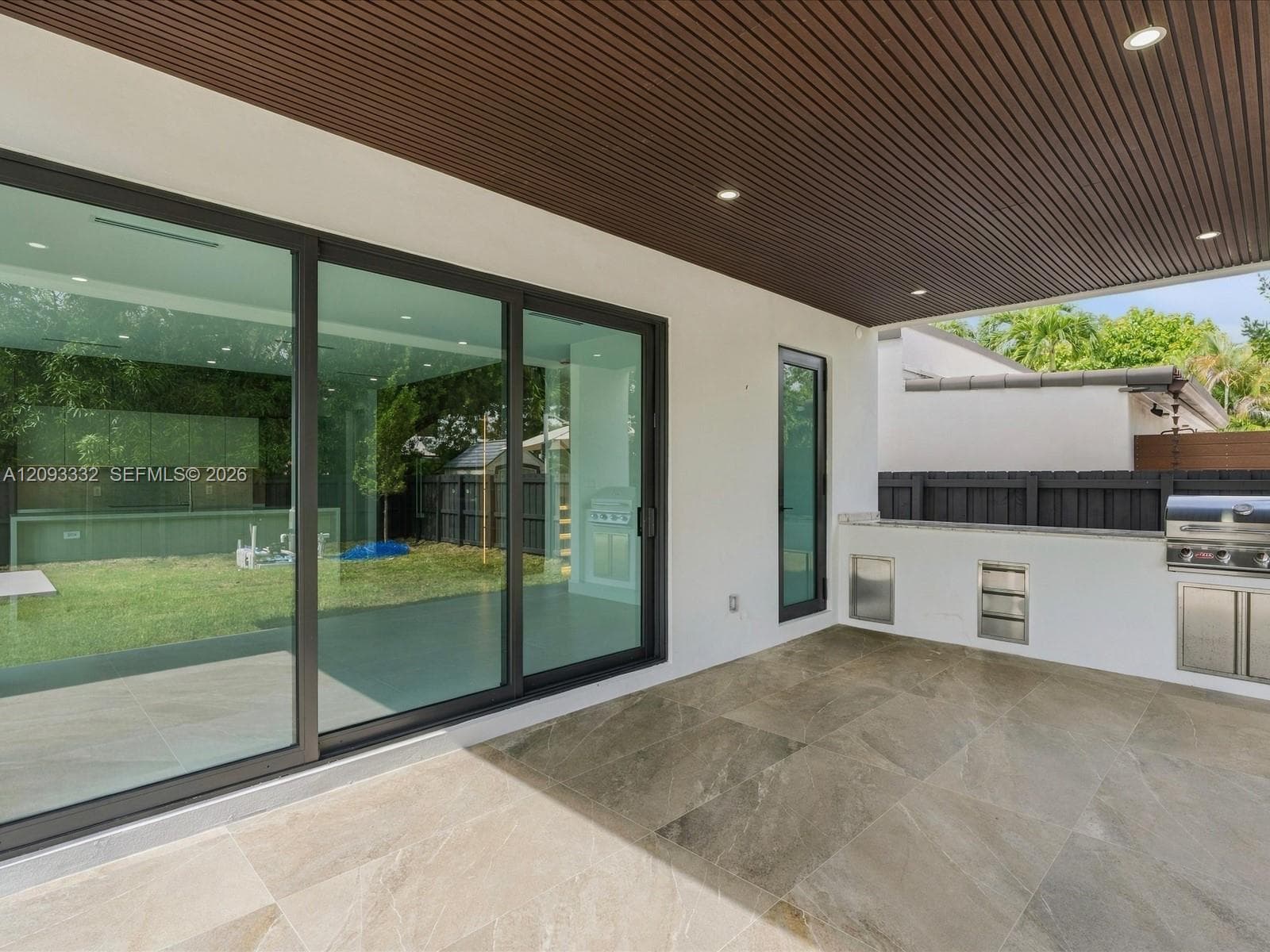 Covered patio featuring large-format stone-finish tile flooring, slatted wood-tone ceiling with recessed lighting, multi-panel sliding glass doors, and a built-in outdoor kitchen with stainless steel appliances