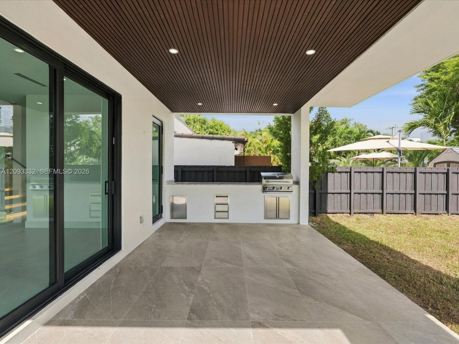 Covered patio featuring large-format stone-finish tile flooring, slatted wood-tone ceiling, recessed lighting, and a built-in outdoor kitchen with a stainless steel grill and stone countertop