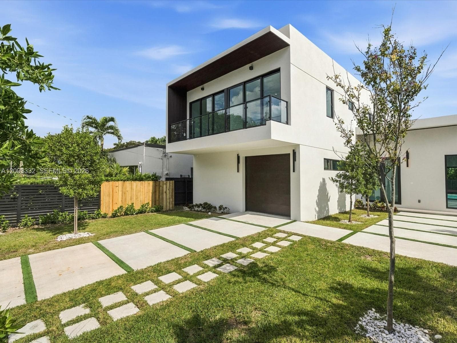 Modern stucco exterior featuring a cantilevered balcony, dark-tinted glass railings, multi-panel sliding doors, and a decorative concrete paver driveway with integrated turf strips
