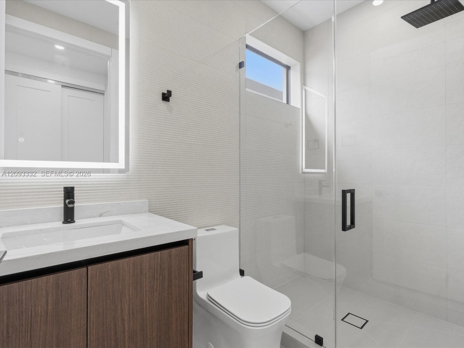 Modern vanity featuring a stone countertop, integrated sink, and illuminated mirror, paired with a glass-enclosed shower with matte black rainfall showerhead and wall-mounted fixtures