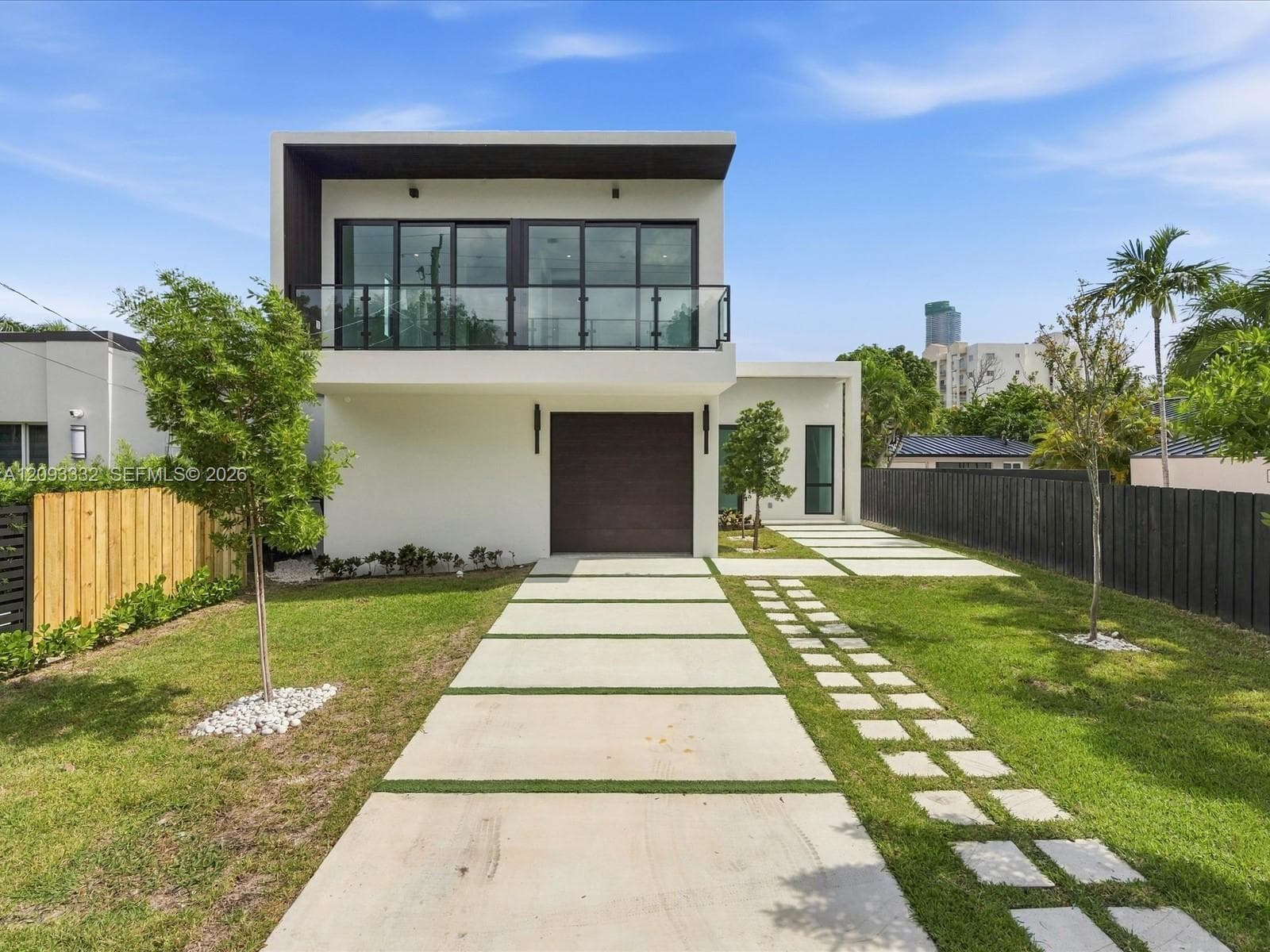 Modern two-story exterior featuring smooth stucco finish, expansive second-level glass balcony with metal railing, single-car garage, and geometric concrete paver driveway