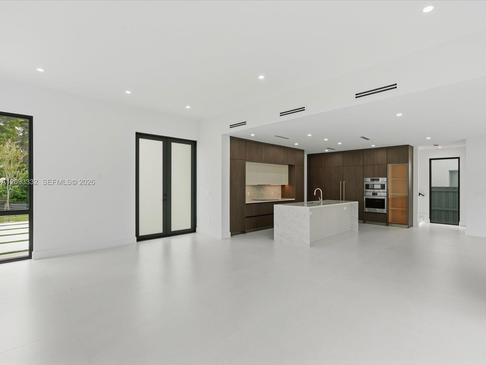 Open-concept interior featuring large-format tile flooring, a kitchen island with a stone-finish countertop, dark wood-finish cabinetry, built-in stainless steel double ovens, and black-framed glass double doors