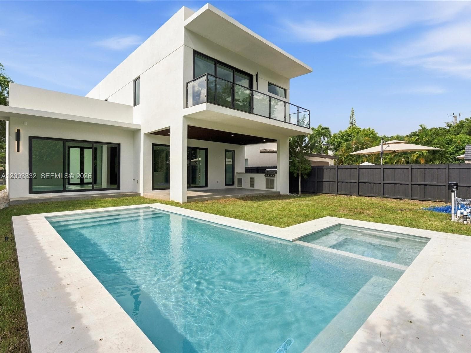 Modern exterior featuring a geometric stucco facade, a second-story balcony with glass railing, a covered patio with built-in outdoor kitchen, and a swimming pool with an attached spa