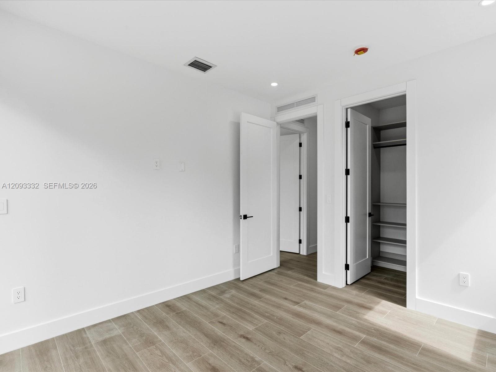 Neutral-toned wood-finish flooring, crisp white walls, minimalist shaker-style interior doors with matte black hardware, and an integrated reach-in closet featuring built-in shelving