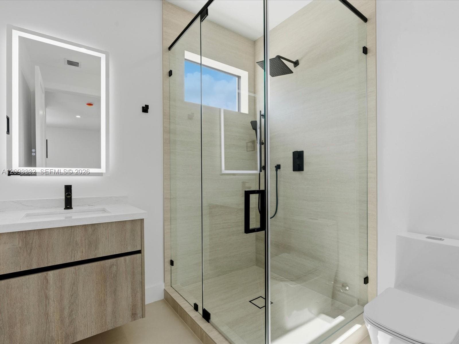 Bathroom featuring a floating wood-finish vanity, illuminated wall mirror, and walk-in shower with floor-to-ceiling tile, glass enclosure, and matte black hardware