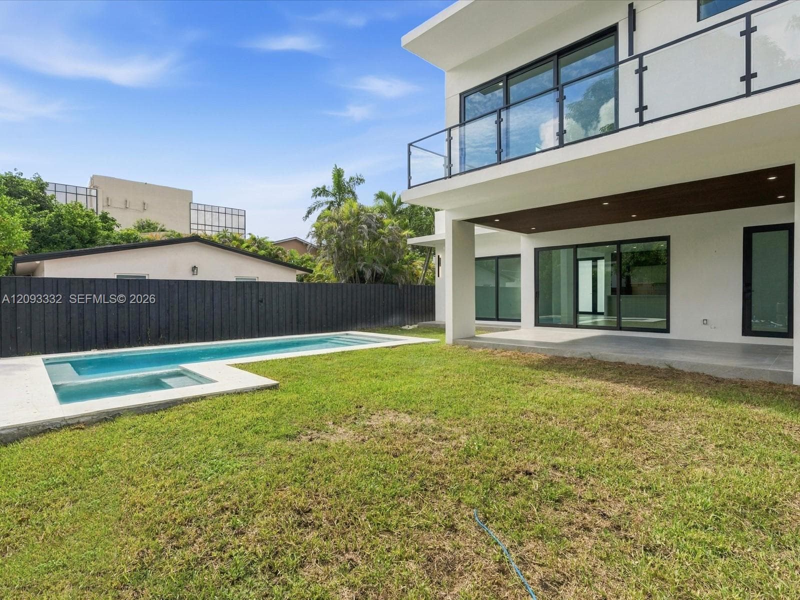 Modern architectural exterior featuring a second-story balcony with glass railings, a covered ground-level patio, an integrated swimming pool with a spa, and a perimeter privacy fence