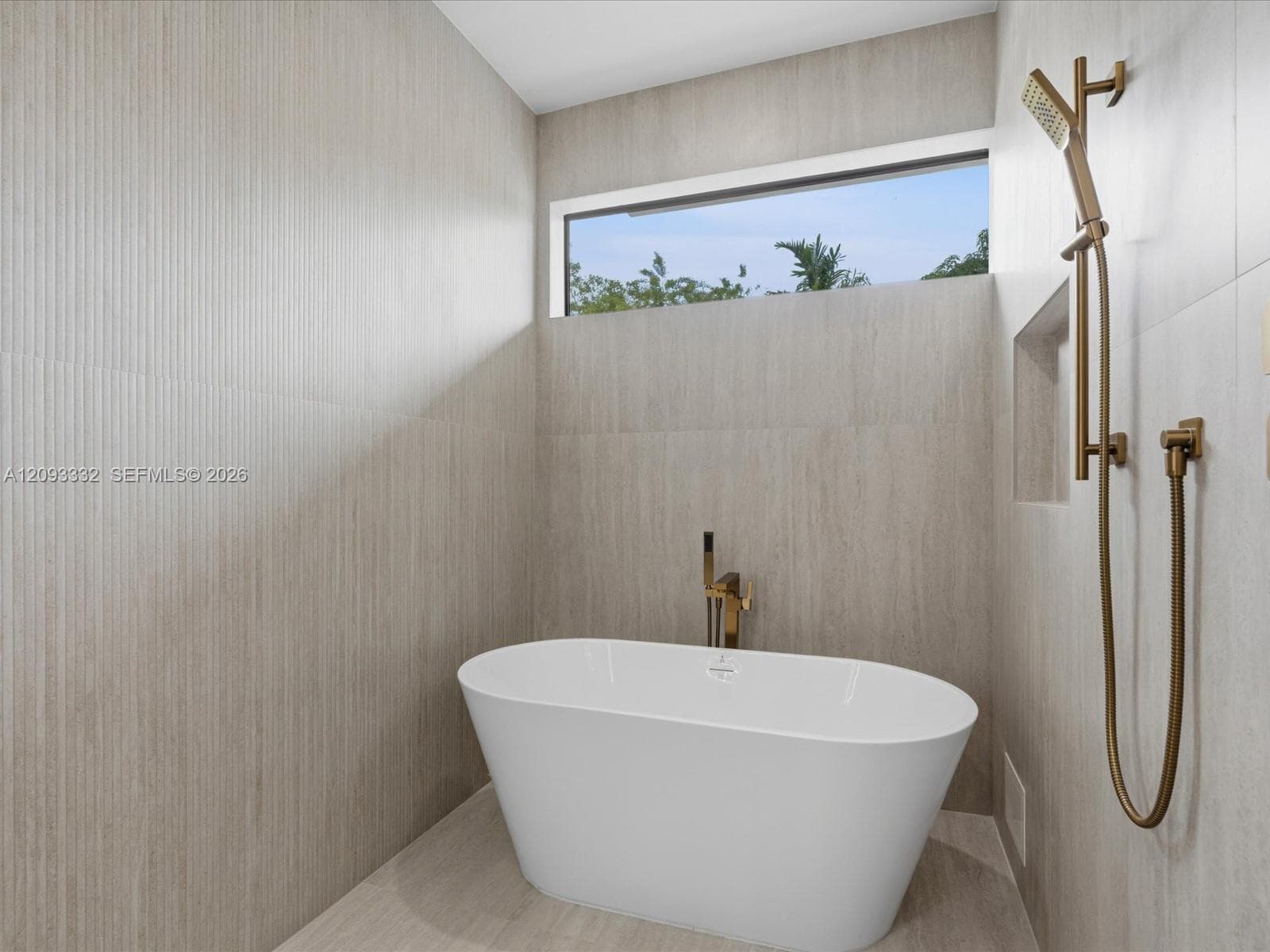 Freestanding soaking tub featuring a metallic bronze floor-mounted faucet, textured stone-finish wall tile, a recessed wall niche, and a metallic bronze handheld shower fixture