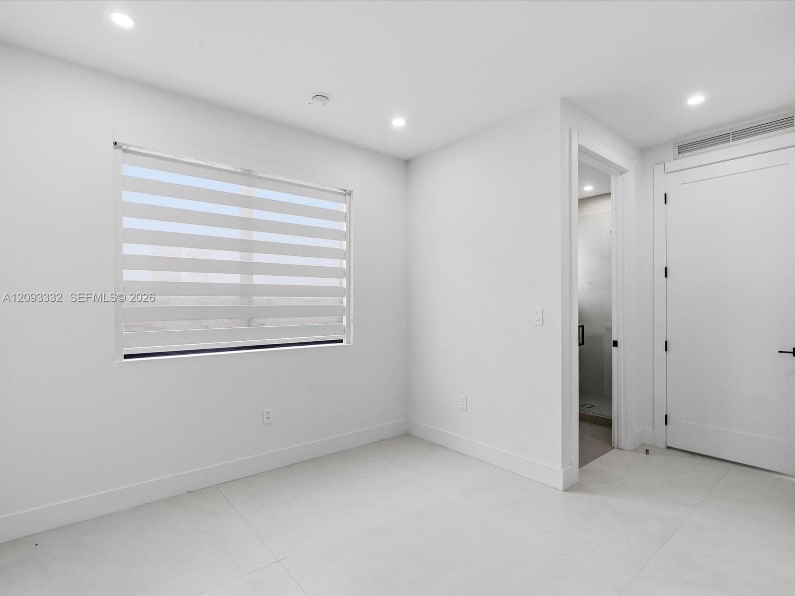 Neutral-toned interior featuring large-format tile flooring, recessed ceiling lighting, a single window with horizontal privacy blinds, and a solid-core interior door with matte black hardware
