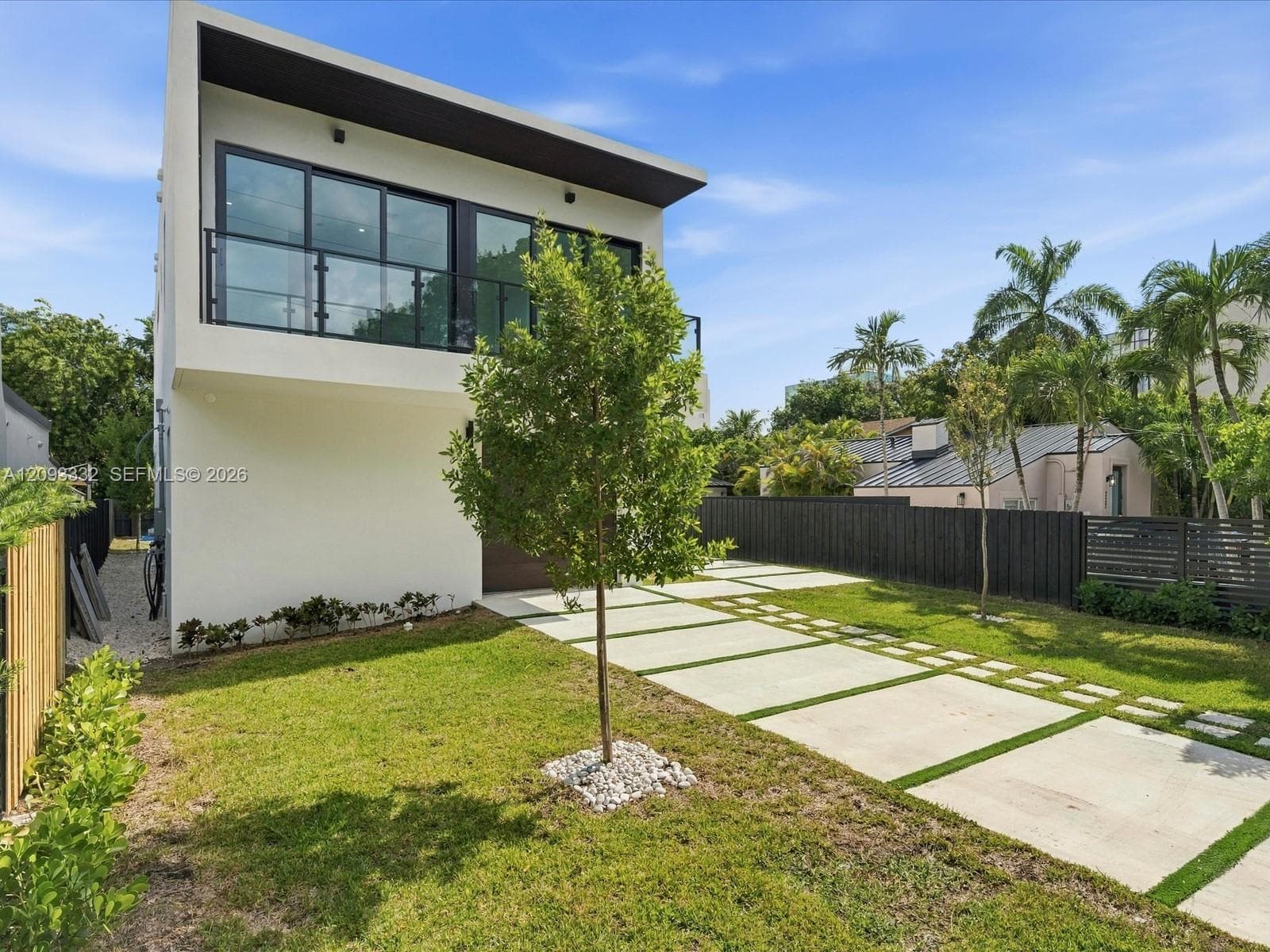 Modern exterior featuring a smooth stucco finish, upper-level balcony with metal railing, flat roofline, oversized sliding glass doors, and a concrete paver driveway with integrated lawn strips