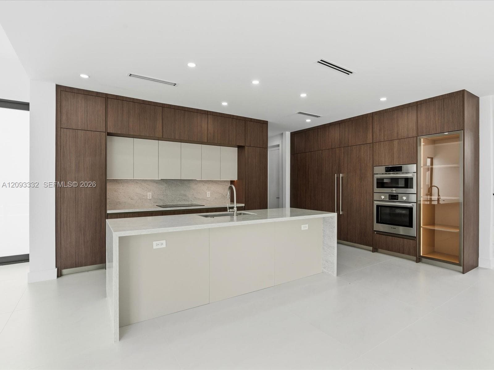Modern kitchen featuring full-height wood-finish cabinetry, a waterfall-edge stone island, built-in stainless steel double ovens, and a glass-front display cabinet