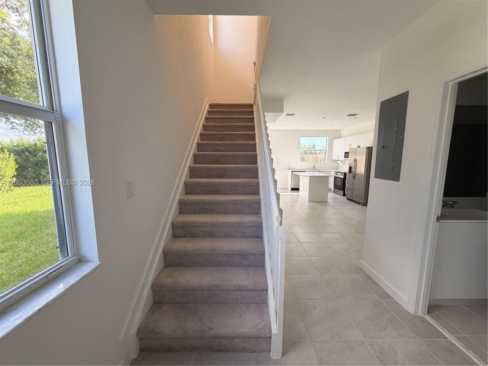 Carpeted staircase with white handrail, large windows, neutral tile flooring, and open-concept kitchen area with central island