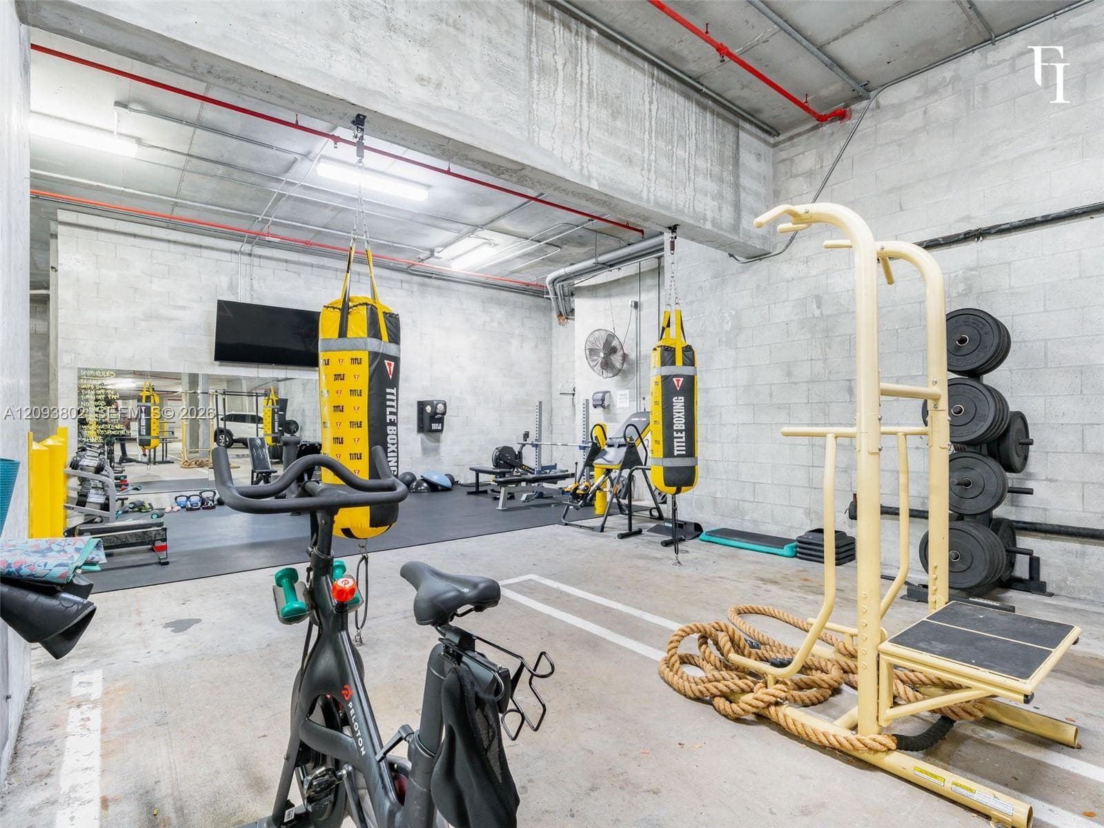 Industrial-style fitness space featuring exposed concrete block walls, high ceilings with exposed piping, large-scale wall mirrors, and recessed overhead fluorescent lighting