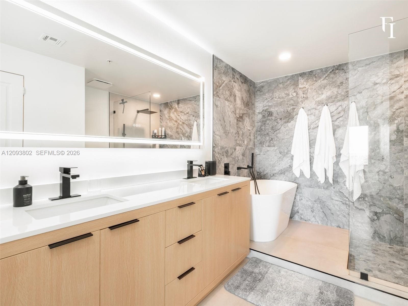 Floating wood-finish vanity with dual integrated sinks and quartz countertops, oversized backlit mirror, freestanding soaking tub, and stone-tiled shower enclosure with glass surround