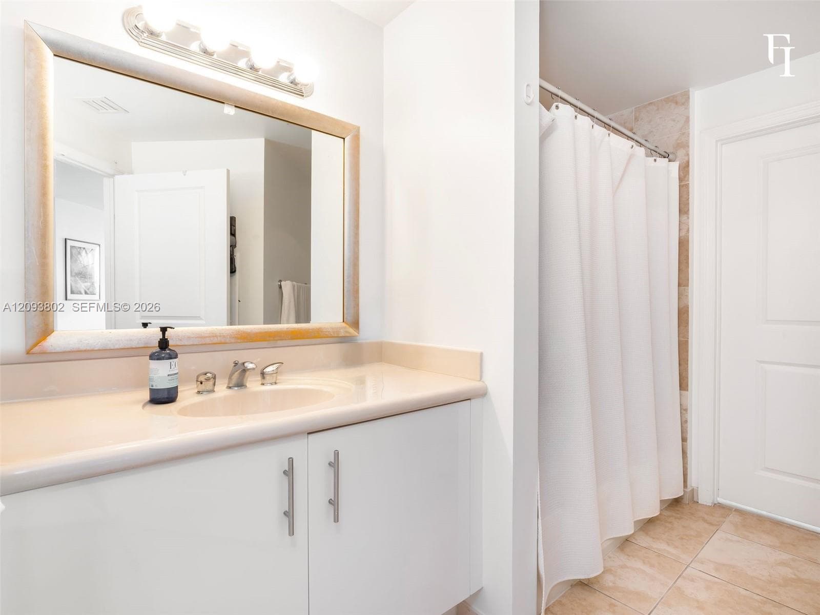 Bathroom featuring a white vanity cabinet with a solid surface countertop, an undermount sink, a framed mirror with integrated lighting, and tile flooring