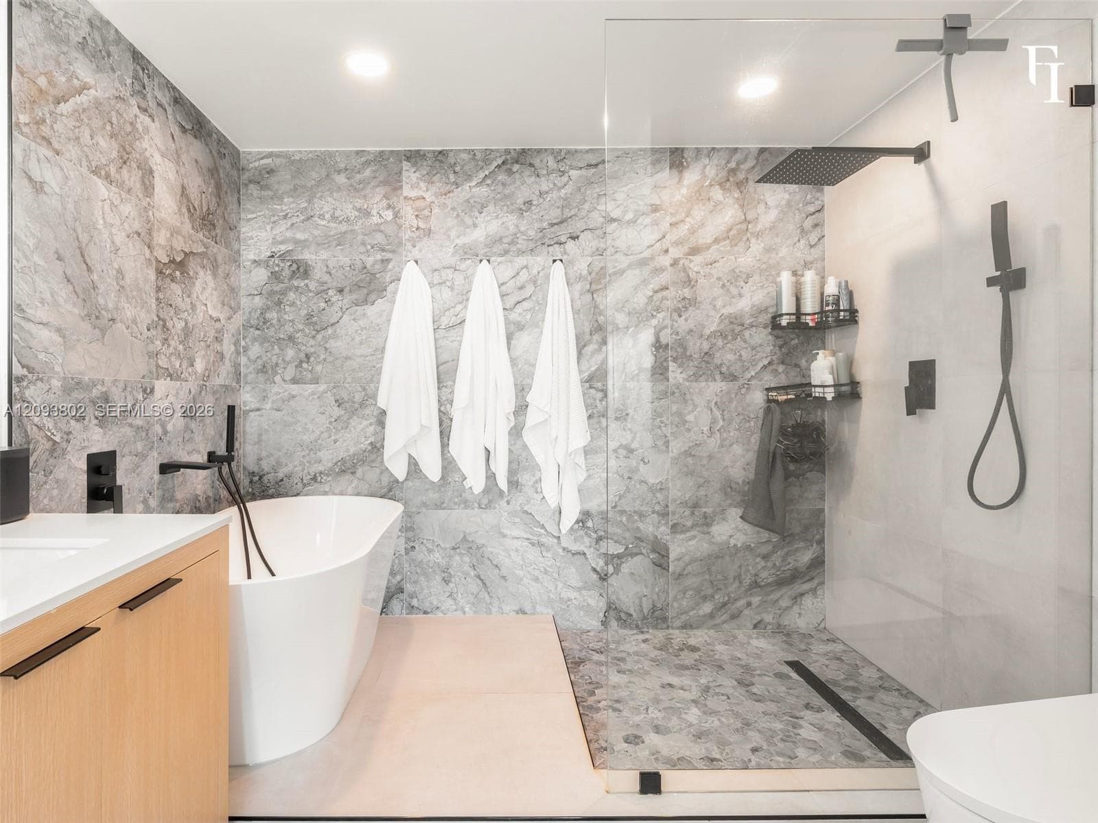 Freestanding soaking tub with matte black wall-mounted faucet, walk-in shower featuring a glass enclosure and rainfall showerhead, and large-format stone-finish tile wall accents