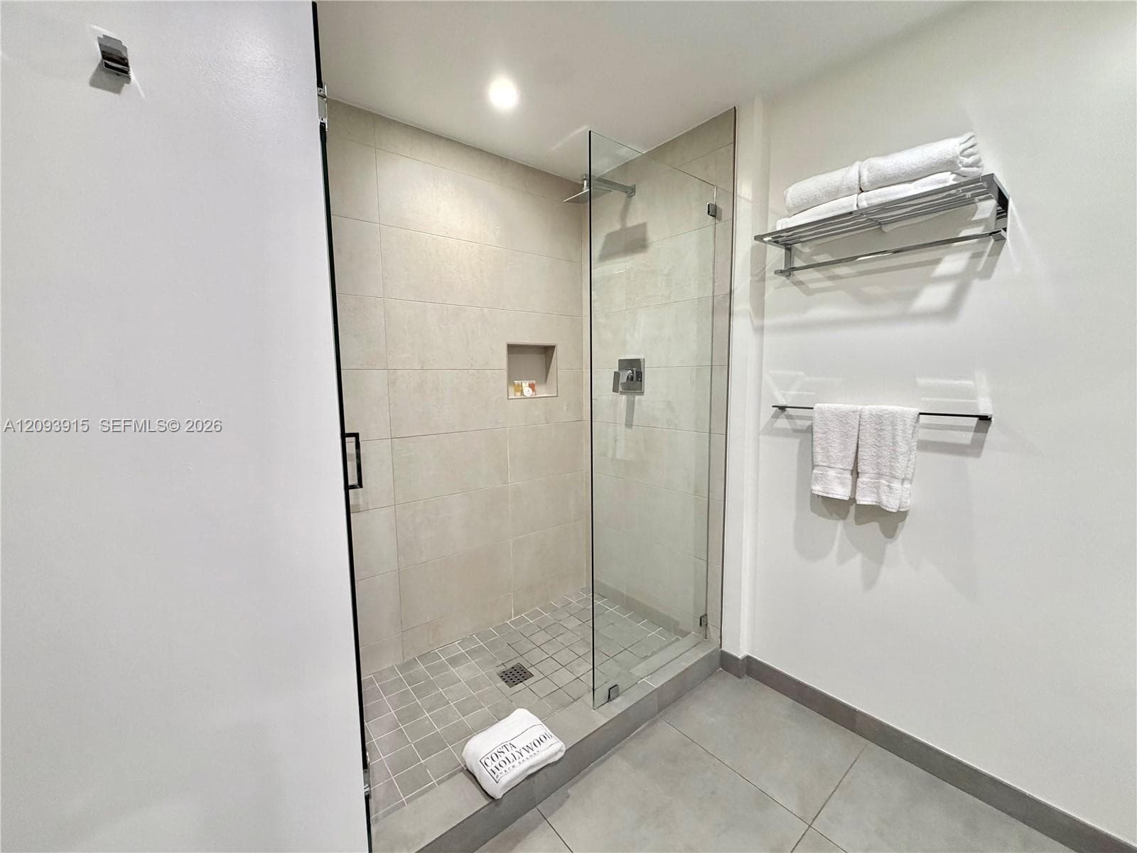 Walk-in shower featuring large-format wall tile, a recessed shampoo niche, a glass enclosure, square mosaic floor tile, and polished chrome fixtures