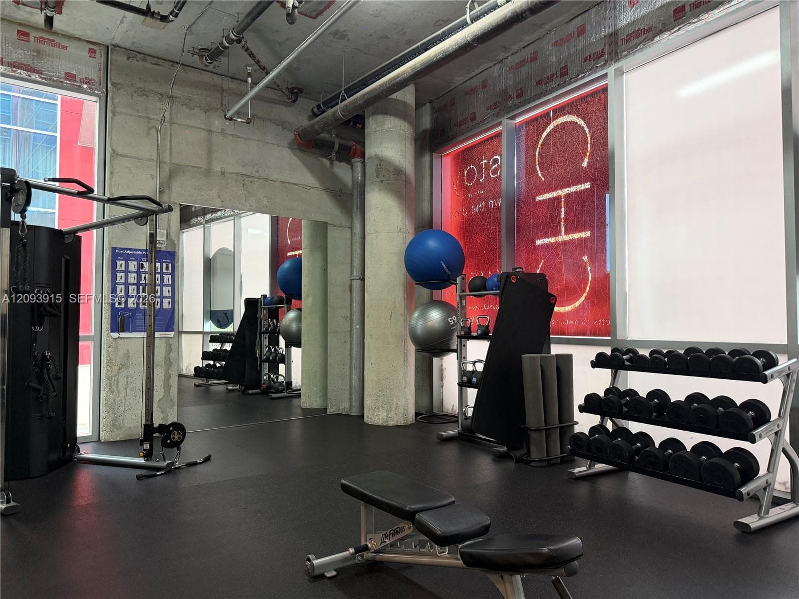 Industrial-style fitness space featuring exposed concrete ceilings, structural pillars, rubberized flooring, and expansive floor-to-ceiling windows