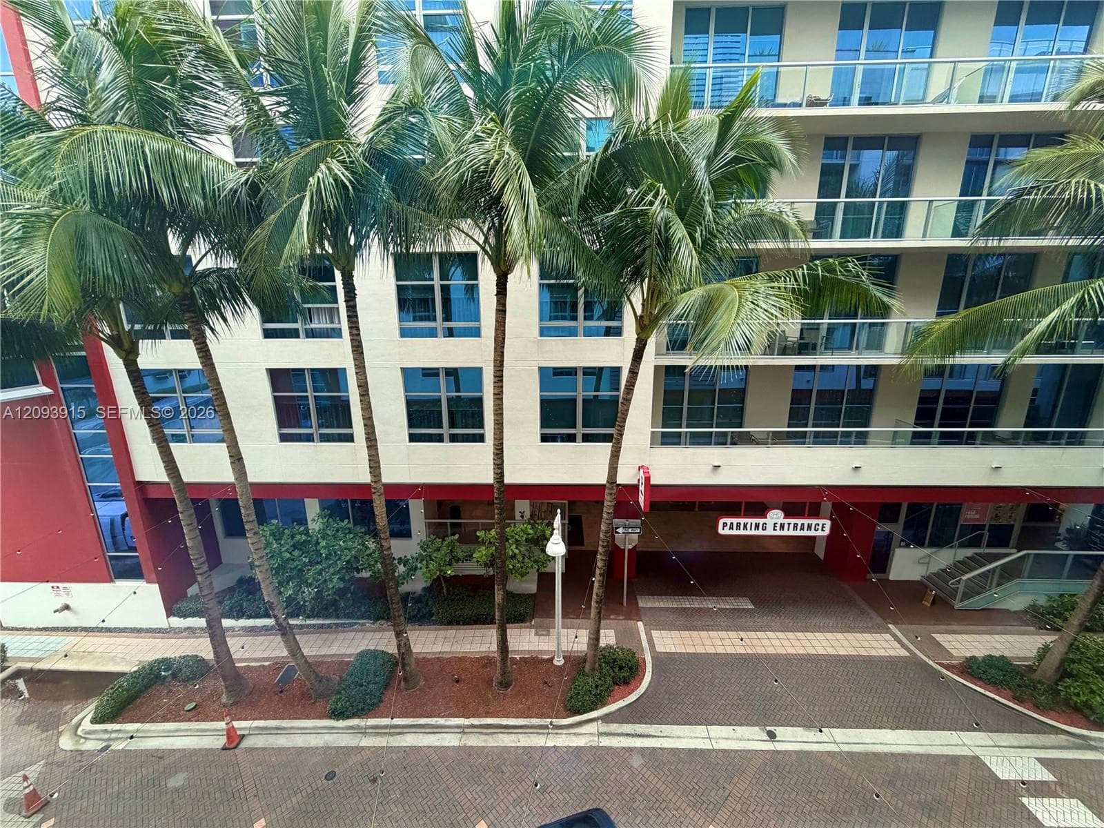 Mid-rise building exterior featuring stucco facade, multi-level glass railed balconies, mature palm trees, and a dedicated parking entrance with paver driveway