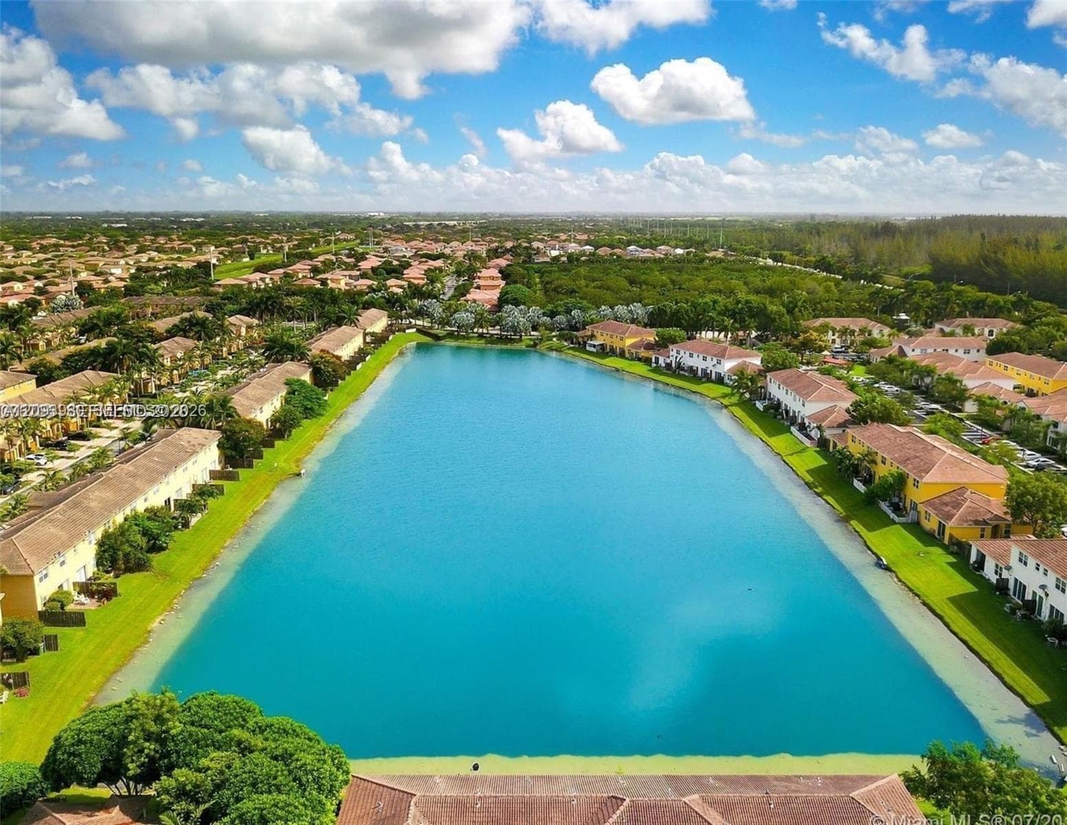 Aerial perspective of a multi-unit residential community centered around an expansive, open-water lake featuring multi-story structures with terracotta-style roofing and manicured green spaces