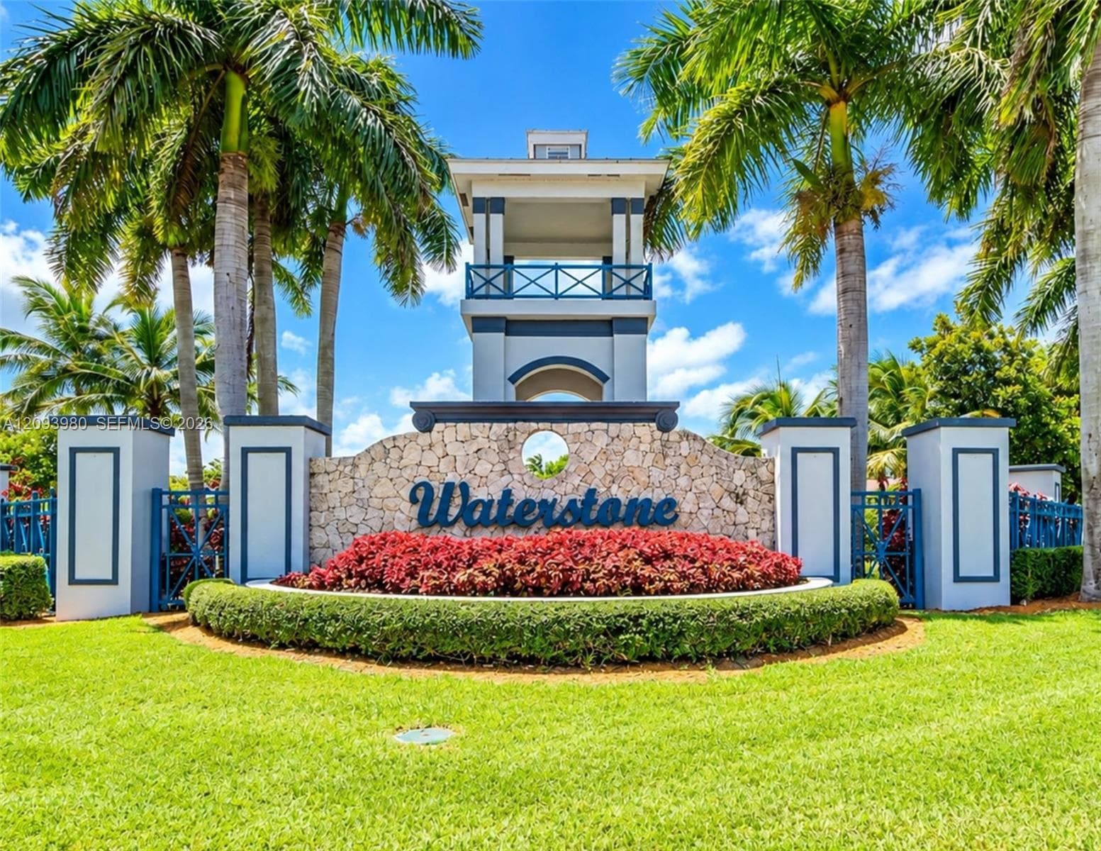 Community entrance featuring a stone masonry facade, tiered tower architectural element, manicured landscaping, and mature palm trees
