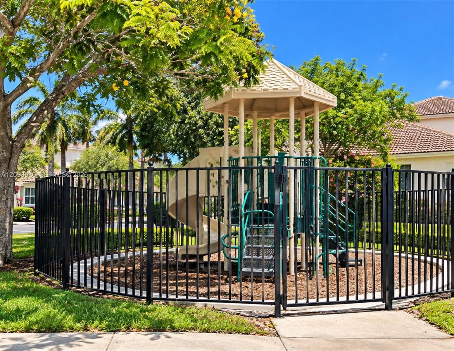 Outdoor recreation area featuring a gazebo-style play structure with a slide, enclosed by black metal fencing and surrounded by mature landscaping