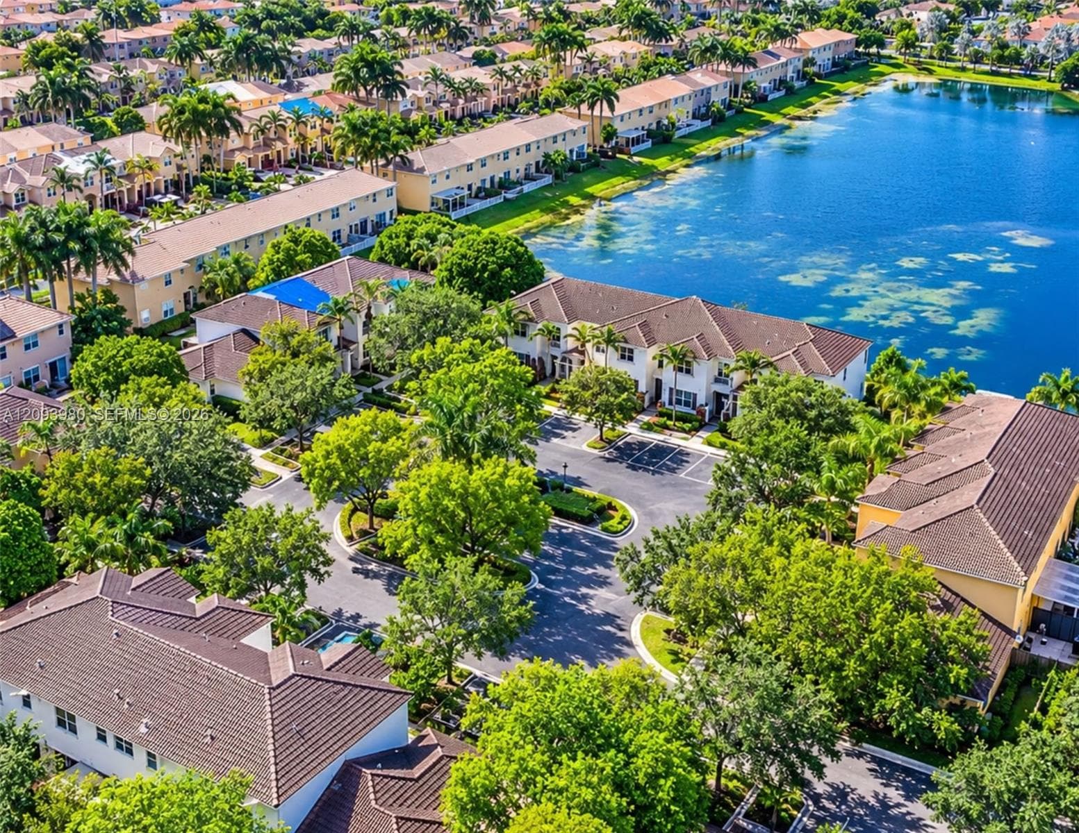 Aerial perspective of a multi-unit residential community featuring clay tile roof systems, mature canopy trees, asphalt parking areas, and waterfront positioning along a lake