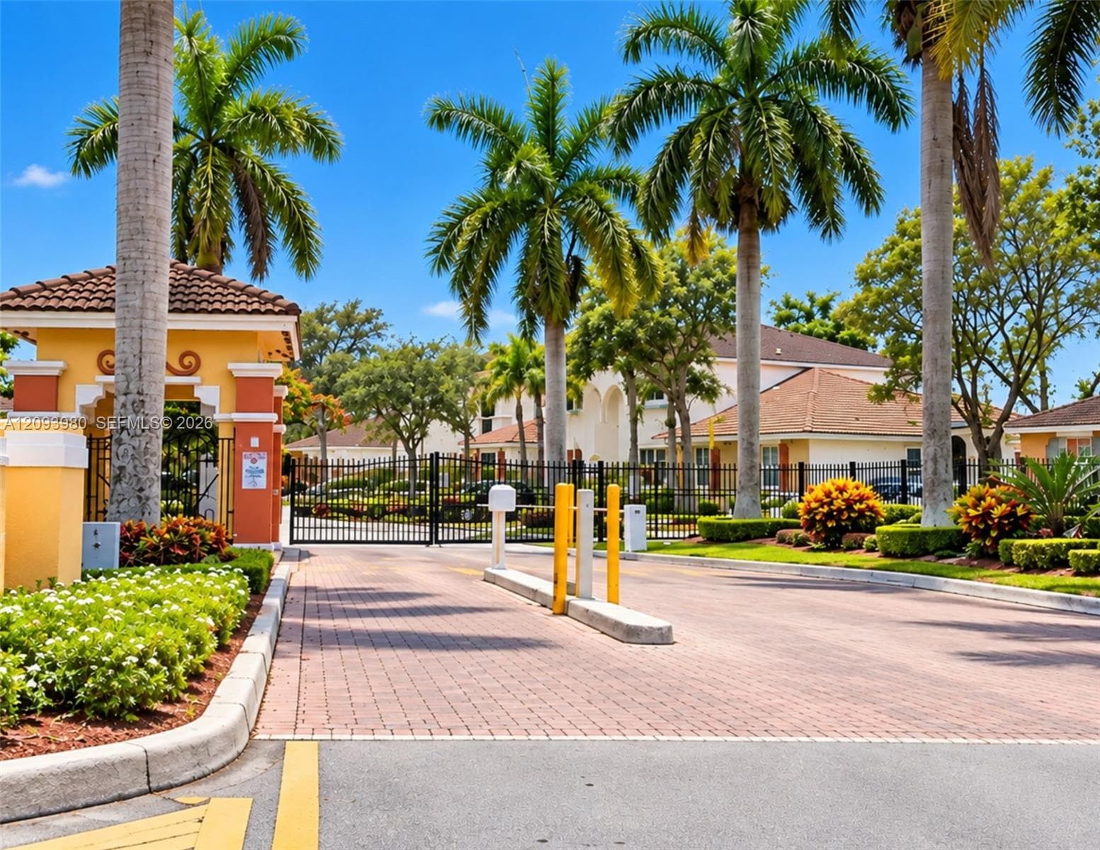Gated entry featuring a stucco security gatehouse, paved brick driveway, perimeter metal fencing, and mature palm landscaping