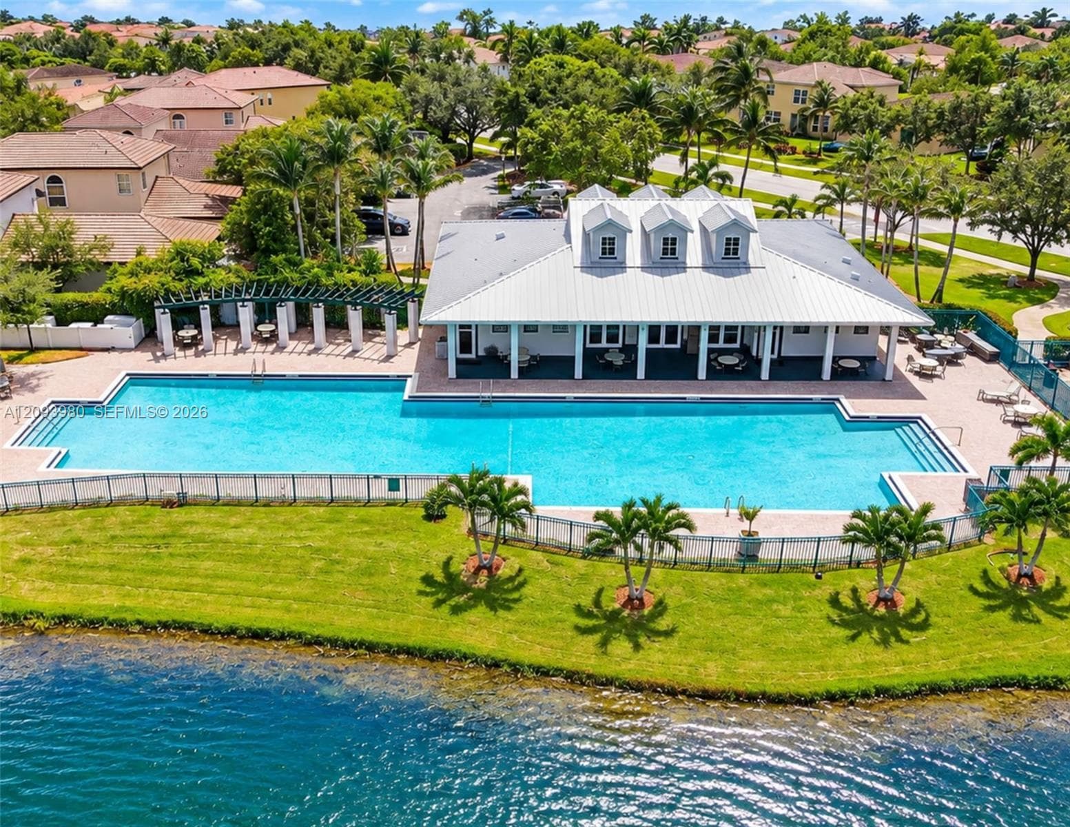 Large lap pool, metal roof pavilion with dormer windows, covered patio featuring a pergola, and manicured lawn situated along a lake