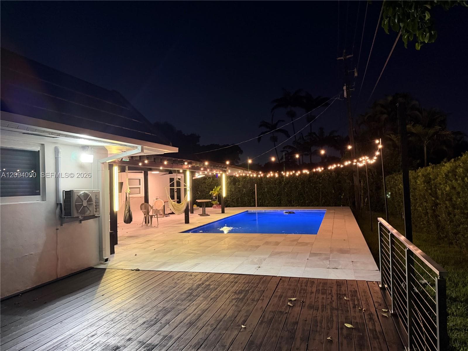 Expansive outdoor living area featuring a rectangular pool, wood-plank decking, and a covered patio with string lighting