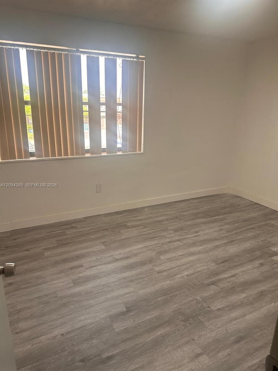 Neutral interior room featuring wood-finish flooring, smooth wall surfaces, and a window with vertical blinds