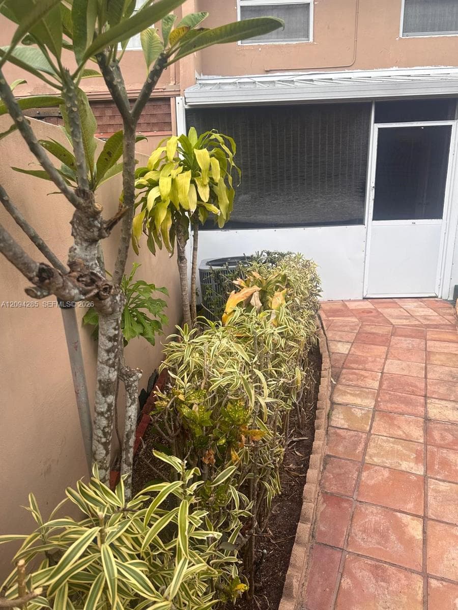 Exterior patio area featuring terracotta-style tile flooring, a screen-enclosed portal, and a landscaped garden bed with mature ornamental plantings
