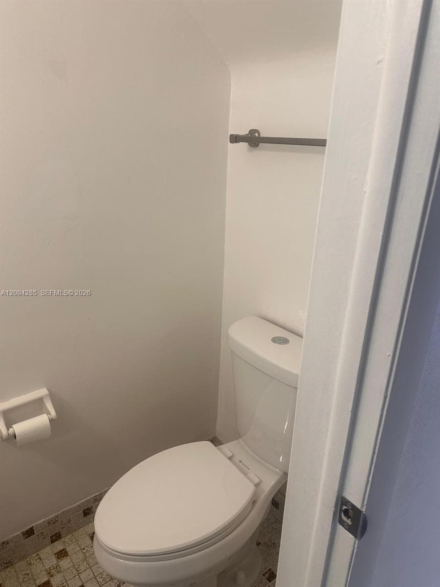 Two-piece toilet with a push-button flush mechanism, wall-mounted metal towel bar, fixed toilet paper holder, and mosaic tile flooring