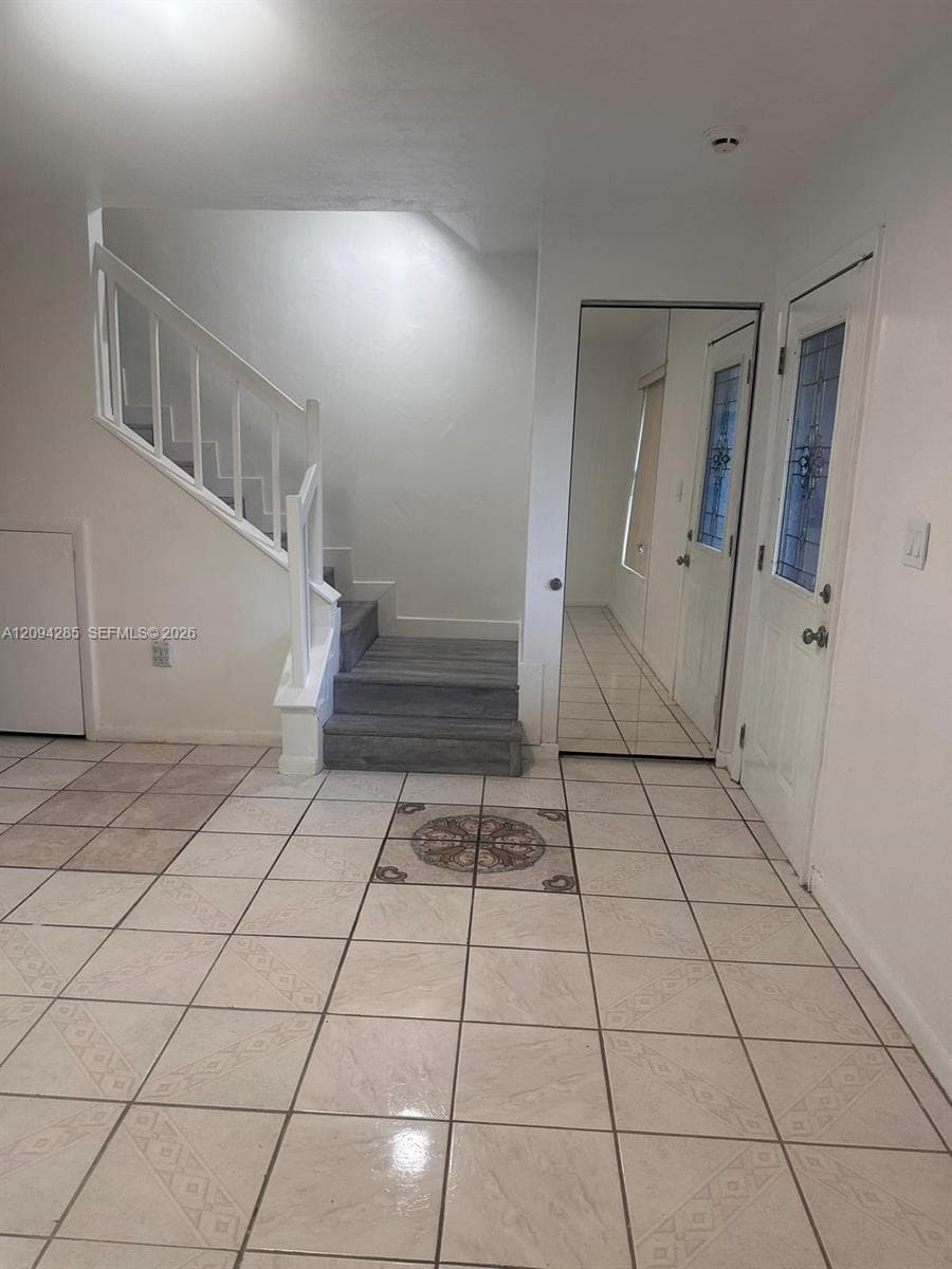 Foyer featuring ceramic tile flooring with decorative medallion inlay, white staircase with wooden balusters, mirrored closet doors, and an exterior door with stained glass insert