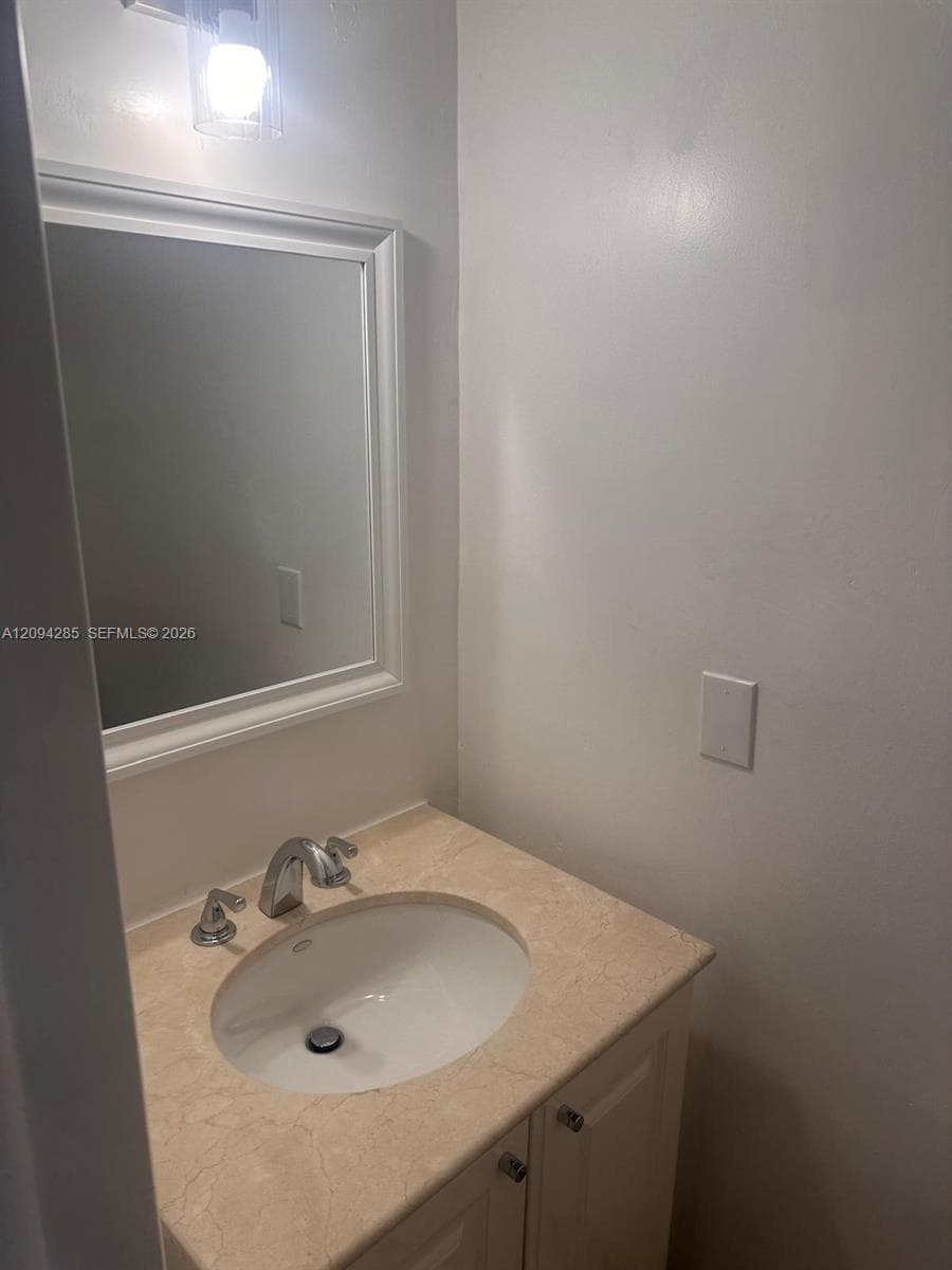 Neutral-toned stone vanity countertop featuring an integrated undermount sink, dual-handle polished chrome faucet, white cabinetry with silver hardware, and a framed wall mirror with a single-light wall-mounted fixture