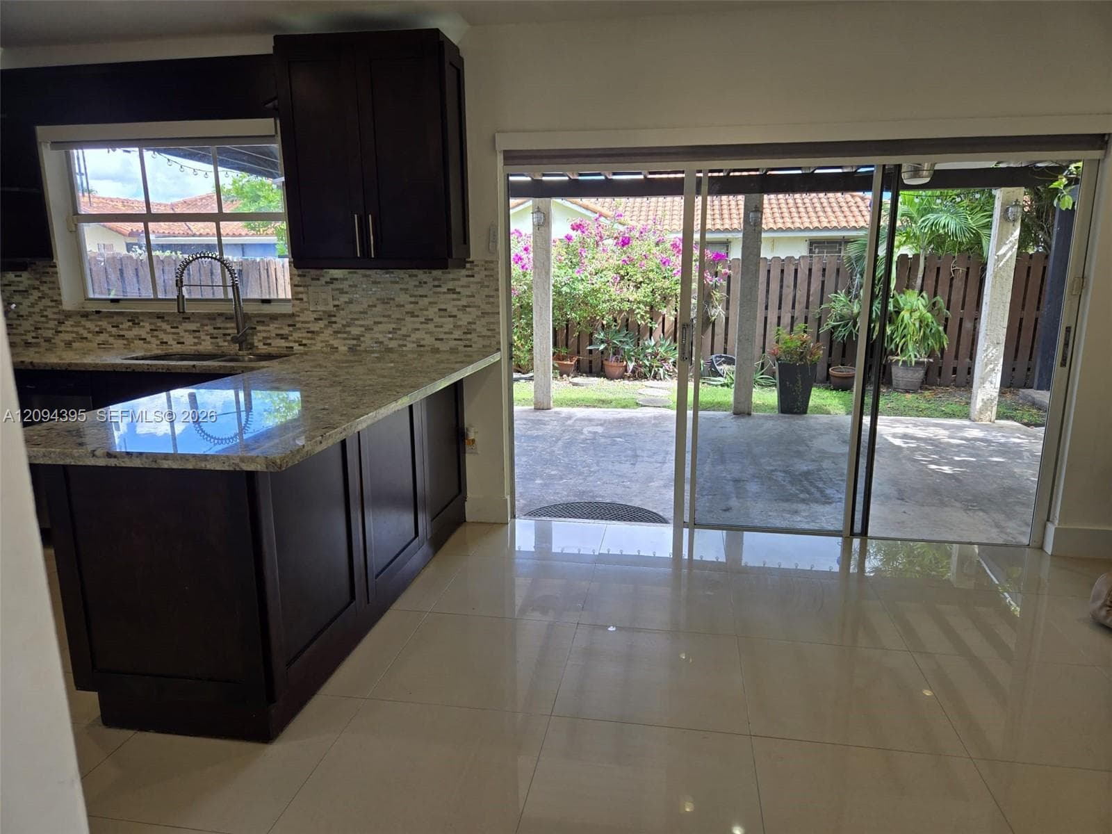 Kitchen featuring dark wood cabinetry, stone countertops, a mosaic tile backsplash, polished tile flooring, and sliding glass doors opening to a covered patio