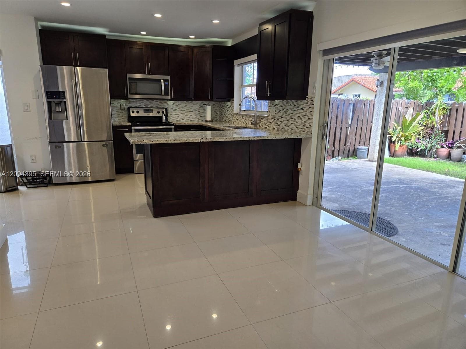 Kitchen featuring dark wood cabinetry, stone-surface countertops, mosaic tile backsplash, stainless steel appliances, and large-format tile flooring