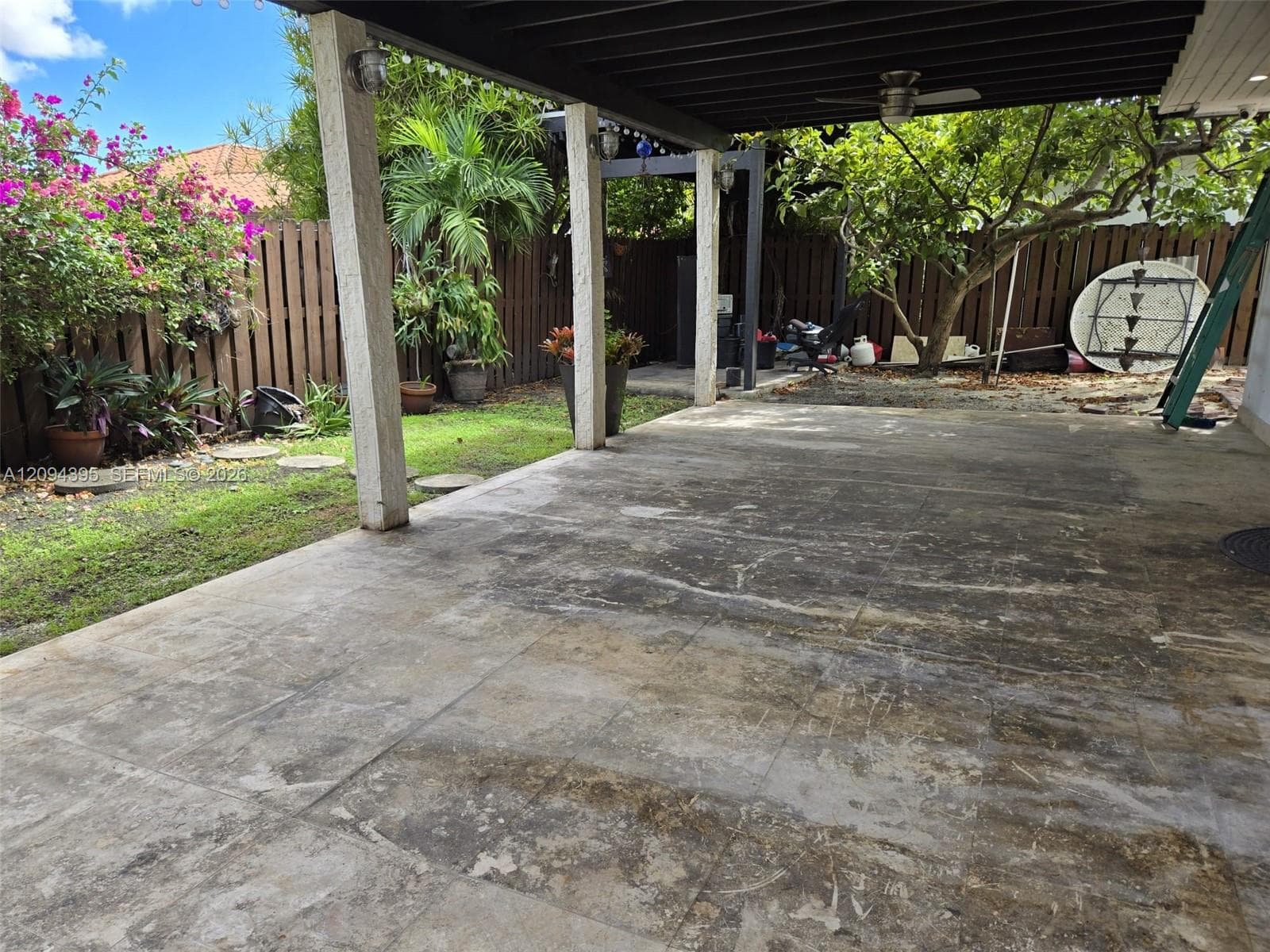 Covered patio with a beamed ceiling, stone-finish flooring, and supporting columns, complemented by a fenced backyard featuring mature trees and established landscaping