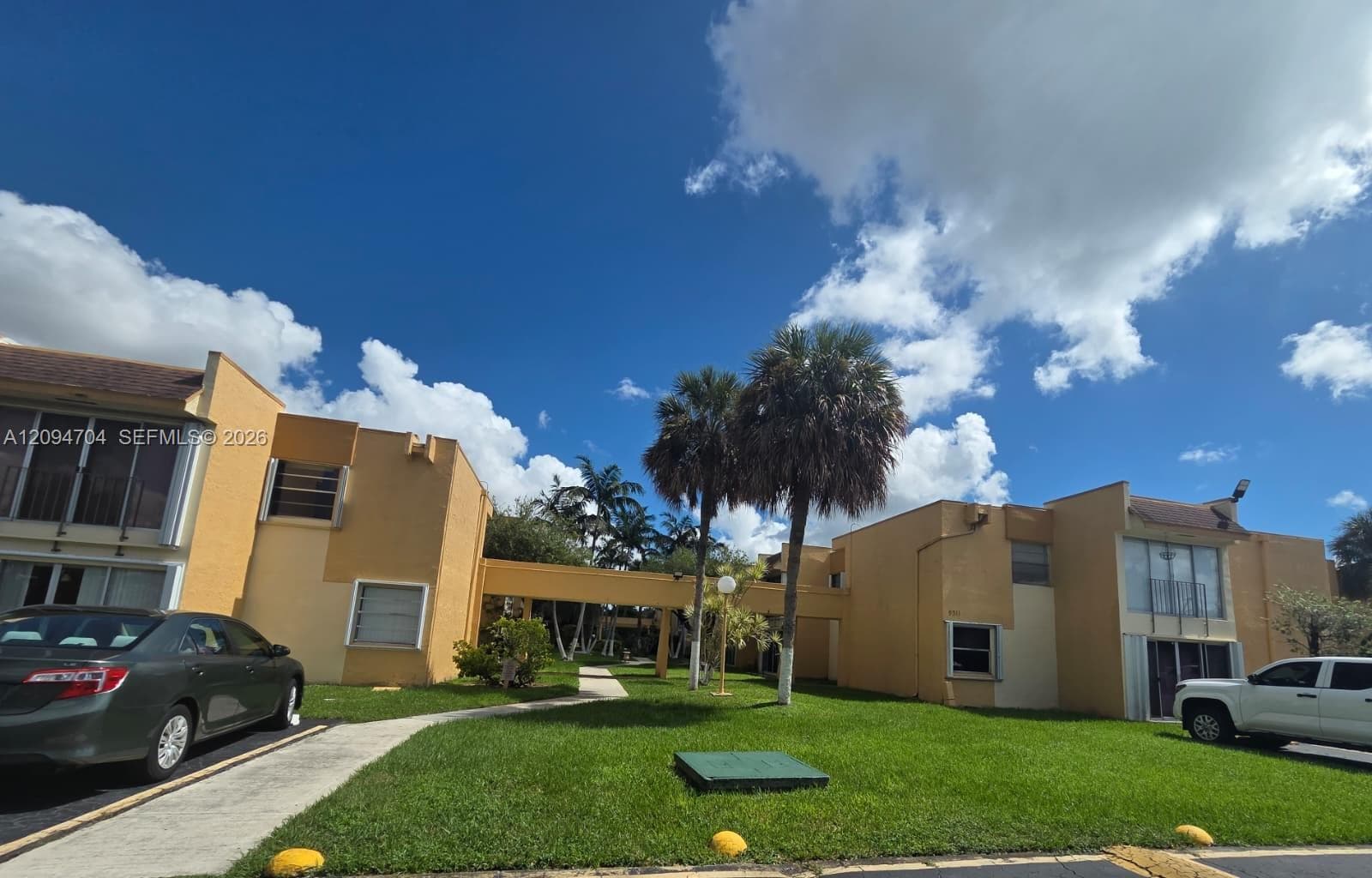 Two-story exterior with stucco facade, covered walkway, balcony access, and mature palm trees