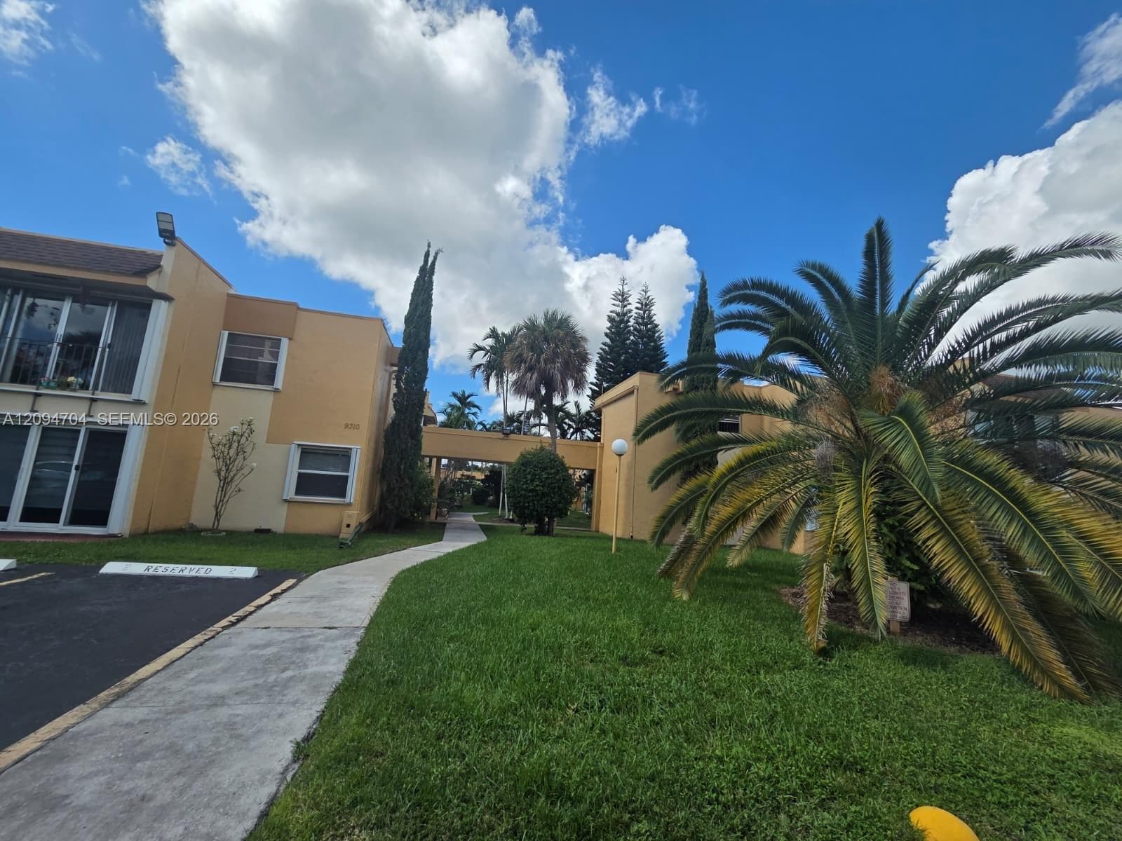 Two-story exterior with stucco finish, elevated covered walkway, and manicured landscaping featuring mature palm trees and green space