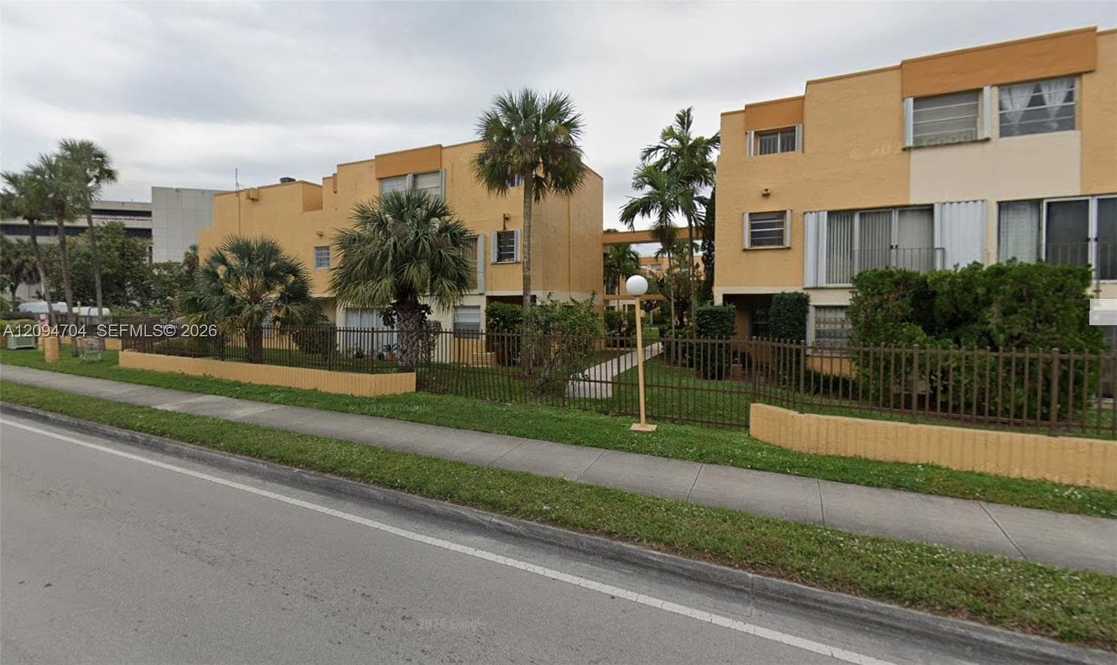 Multi-story masonry exterior featuring a stucco finish, metallic perimeter fencing, and mature palm landscaping