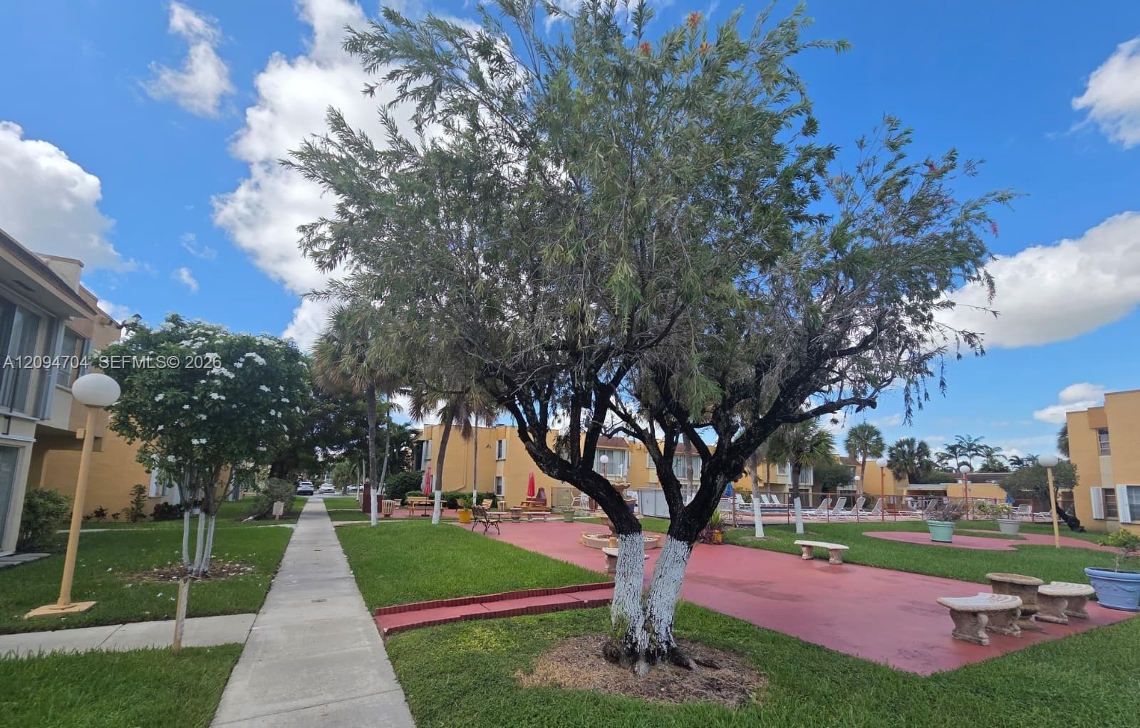 Common area featuring mature landscaping, paved pathways, concrete benches, and stucco building exteriors
